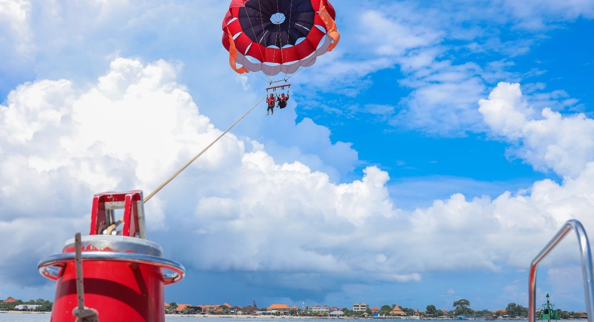 Two people parasailing over the ocean under a red and black parachute, with a clear blue sky filled with fluffy clouds in the background and a winch boat foreground, offering an exciting water sports activity